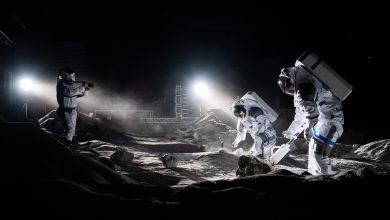 ESA astronauts Matthias Maurer and Aidan Cowley work in a simulated moon landscape, with stage lights recreating the lighting on the lunar surface. Another photographer dressed in protective gear stands to the left.