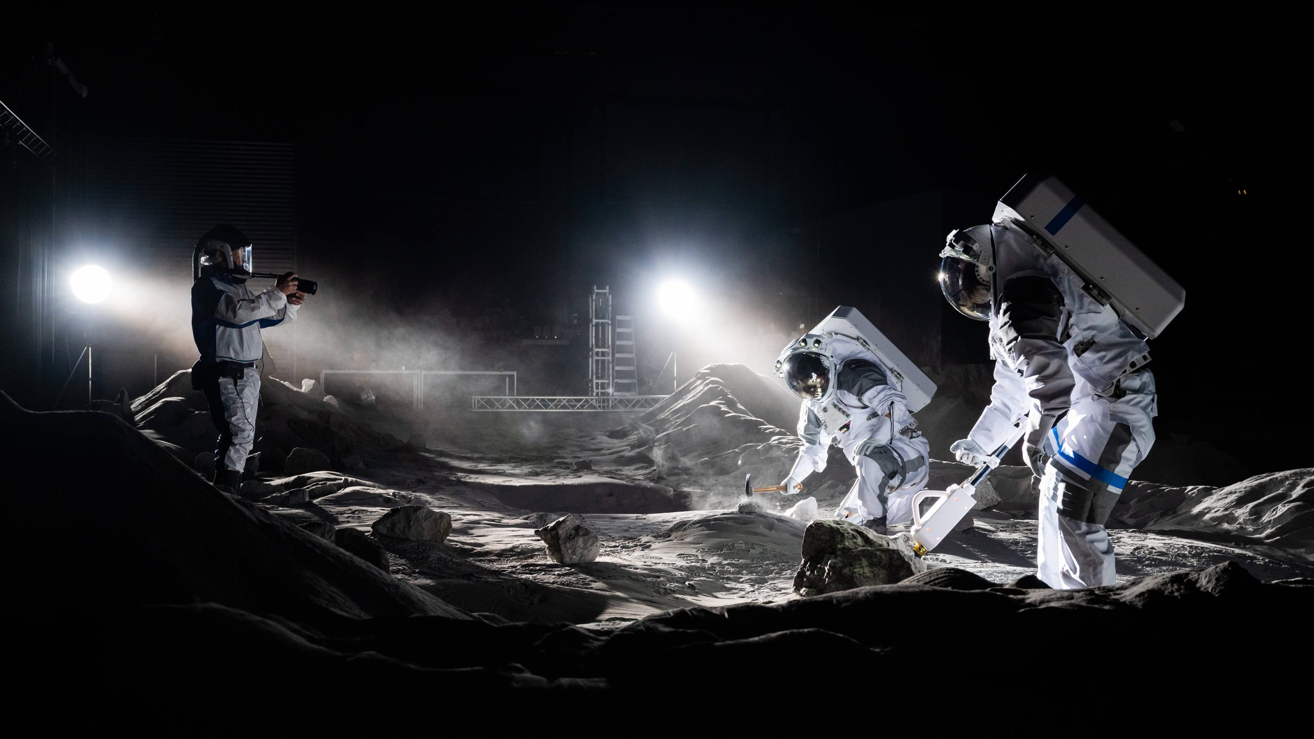 ESA astronauts Matthias Maurer and Aidan Cowley work in a simulated moon landscape, with stage lights recreating the lighting on the lunar surface. Another photographer dressed in protective gear stands to the left.